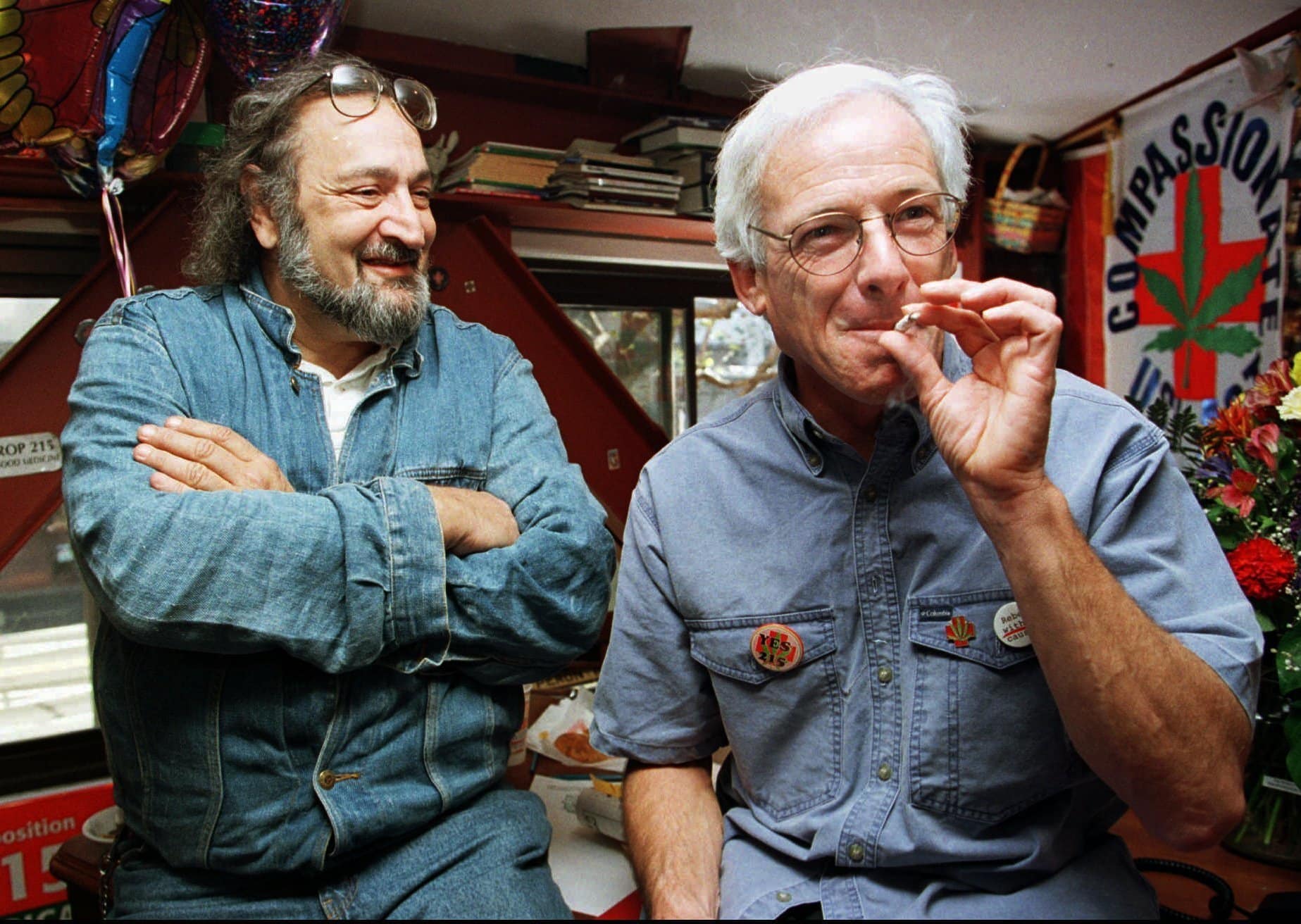 Jack Herer, renowned cannabis activist and author, stands smiling on the left in a denim shirt, while Dennis Peron, pioneer of the medical marijuana movement, sits on the right, smoking a joint. A "Compassionate Use Act" banner hangs in the background.
