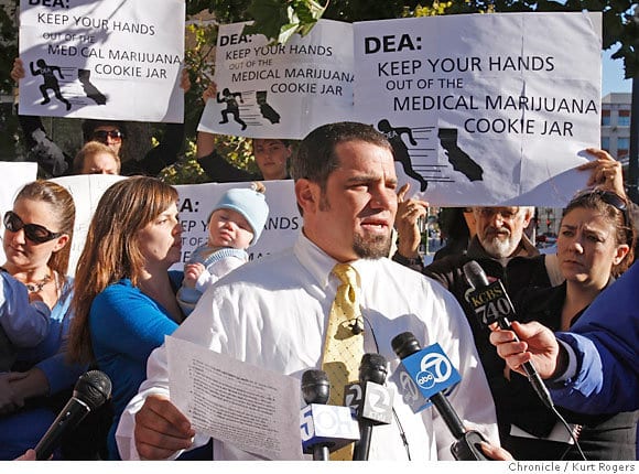 Mickey Martin speaks at a press conference. He is surrounded by supporters. They hold signs that protest DEA interference with medical marijuana. One sign reads, "DEA: Keep Your Hands Out of the Medical Marijuana Cookie Jar."