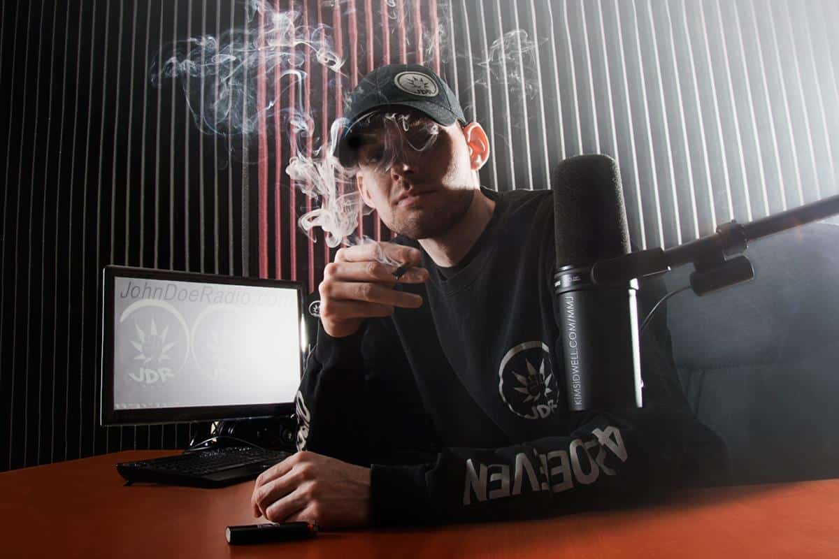 Tim Martin sits at a broadcasting desk. Smoke curls around him. He wears a cap and a long-sleeve shirt with the John Doe Radio logo. A microphone and computer are in the background.