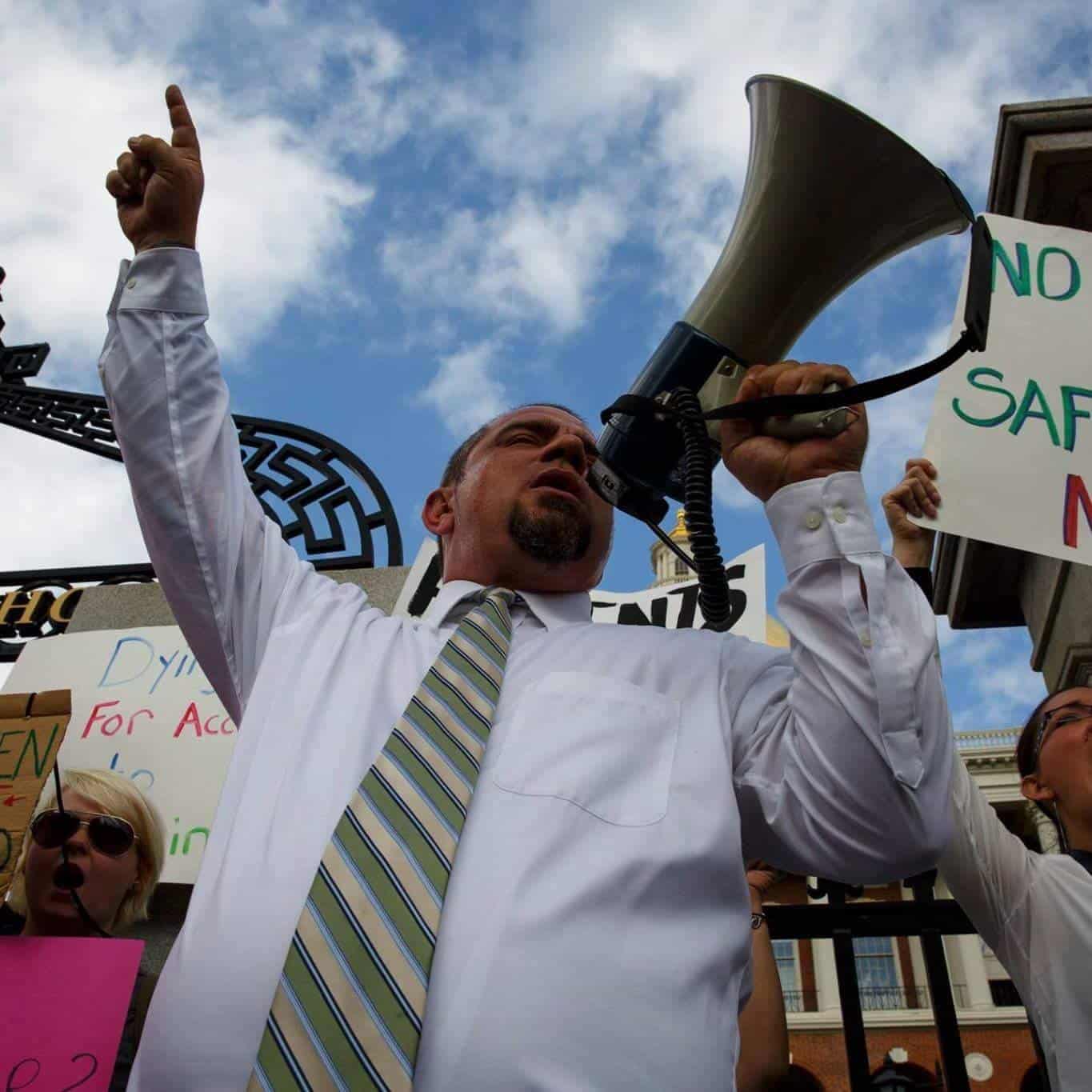 Legendary cannabis activist Mickey Martin passionately addresses a crowd during a protest, holding a megaphone in one hand and raising the other to emphasize his message. Supporters around him hold colorful signs advocating for safe medical access and policy reform.