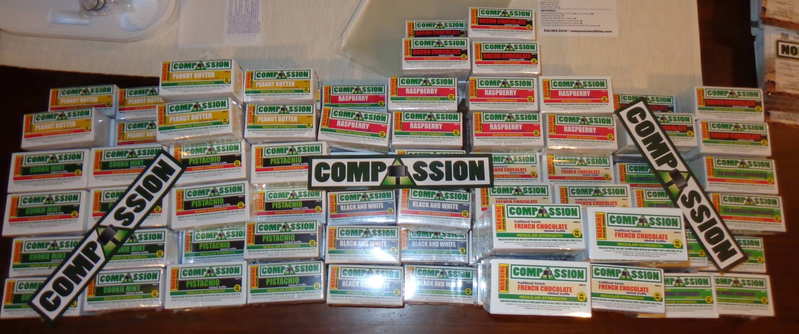 A variety of cannabis edibles from Mickey Martin's Compassion brand is neatly arranged on a table. The display shows different flavors and packaging, with the Compassion logo clearly visible.