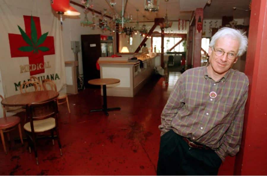 Dennis Peron, pioneer of the medical marijuana movement, leans against a red wall in the interior of a medical cannabis dispensary. A large banner with a cannabis leaf and the words "Medical Marijuana" hangs prominently in the background.