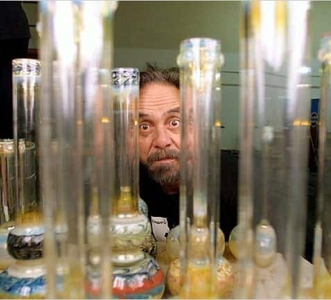 Tommy Chong peers through an array of glass bongs with a curious and playful expression.